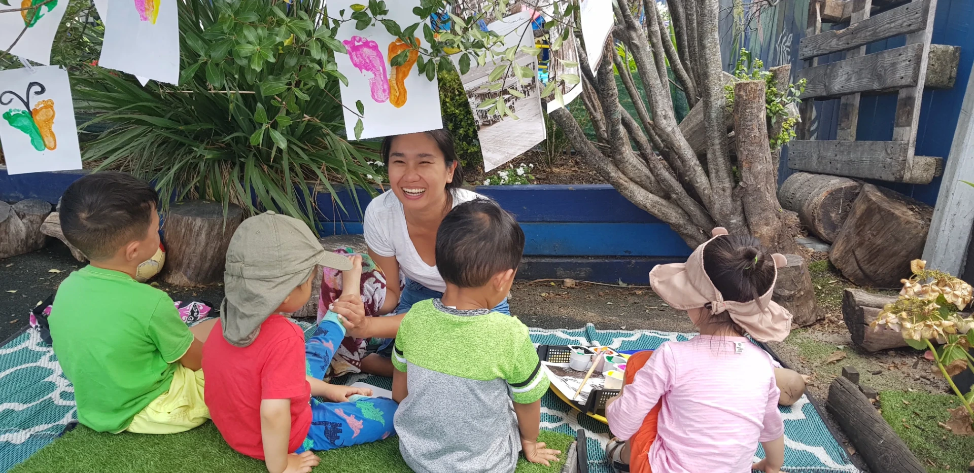 A Growing Minds teacher painting outdoors with a group of children under the trees